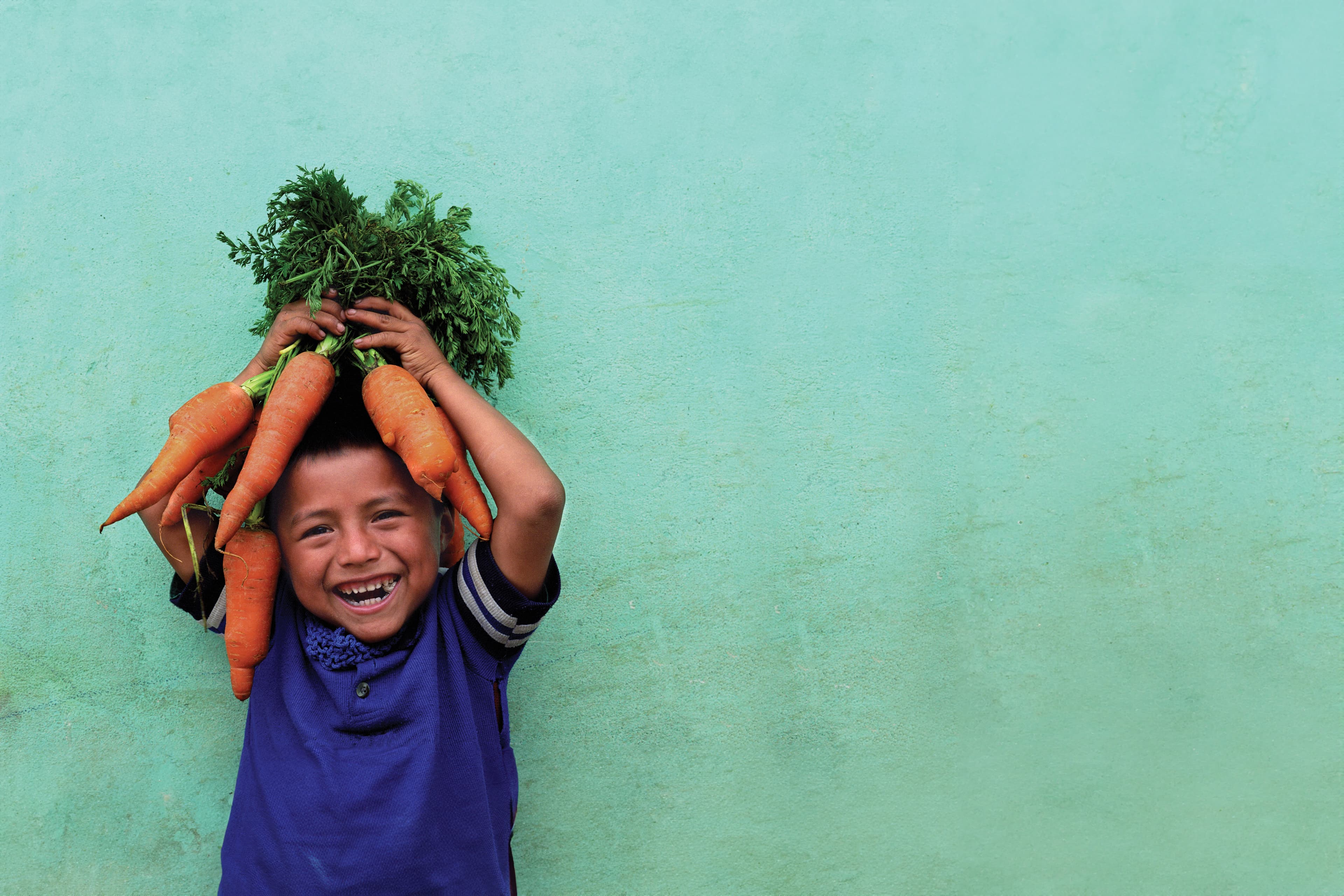A child smiling, holding a large bunch of carrots above his head, against a teal wall